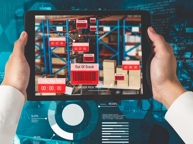 A photo of someone holding a tablet with a picture of boxes sitting on shelves on it. 