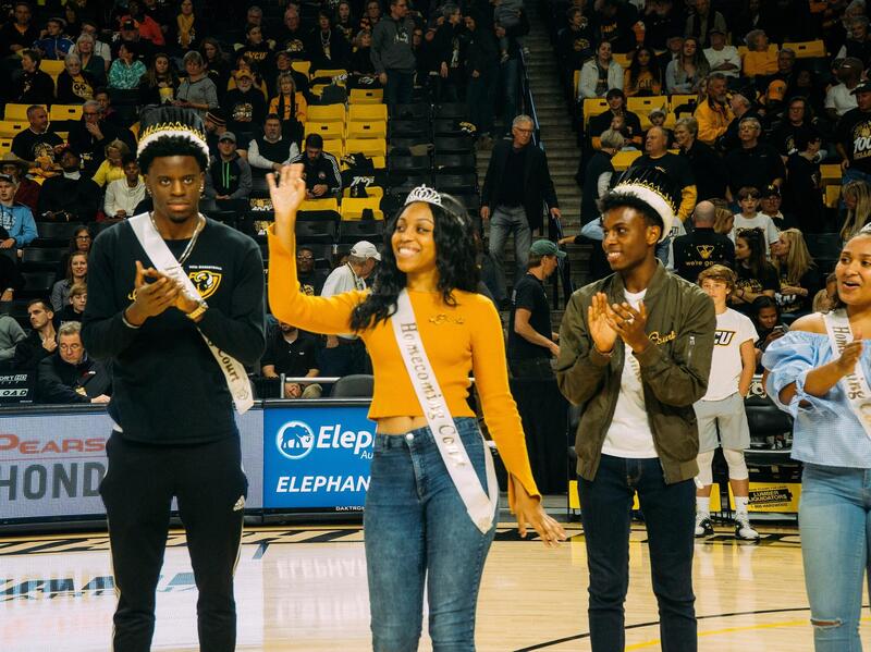Two men and two women wearing crowns and sashes standing on a basketball court. 