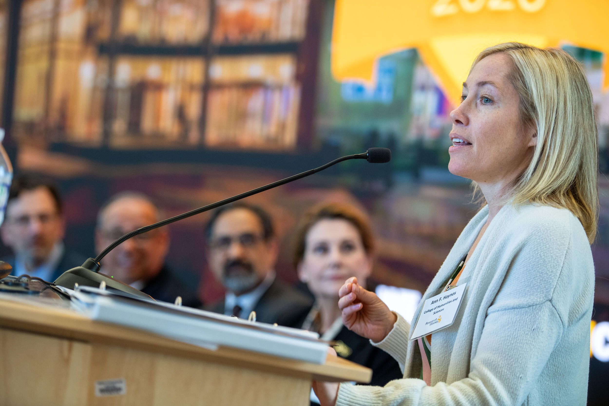 A photo of a woman standing behind a podium. Four people are to the left of her sitting and watching her speak. 