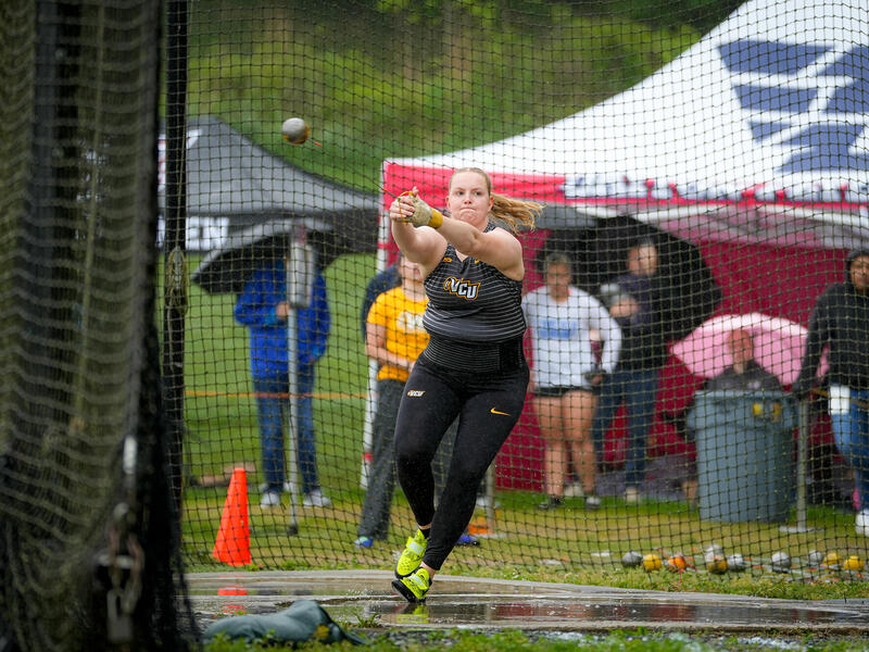 A photo of a woman jumping while holding her arms in front of herself. Behind the woman is a black netted fence with six people standing and sitting behind it. 