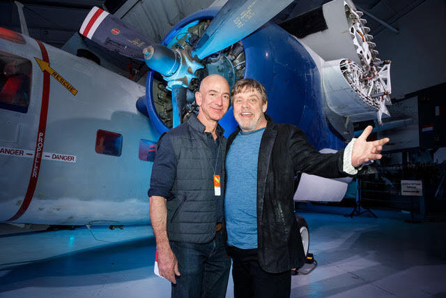 Jeff Bezos (left) and Mark Hamill (right) standing in front of a propeller-driven plane inside of a museum.