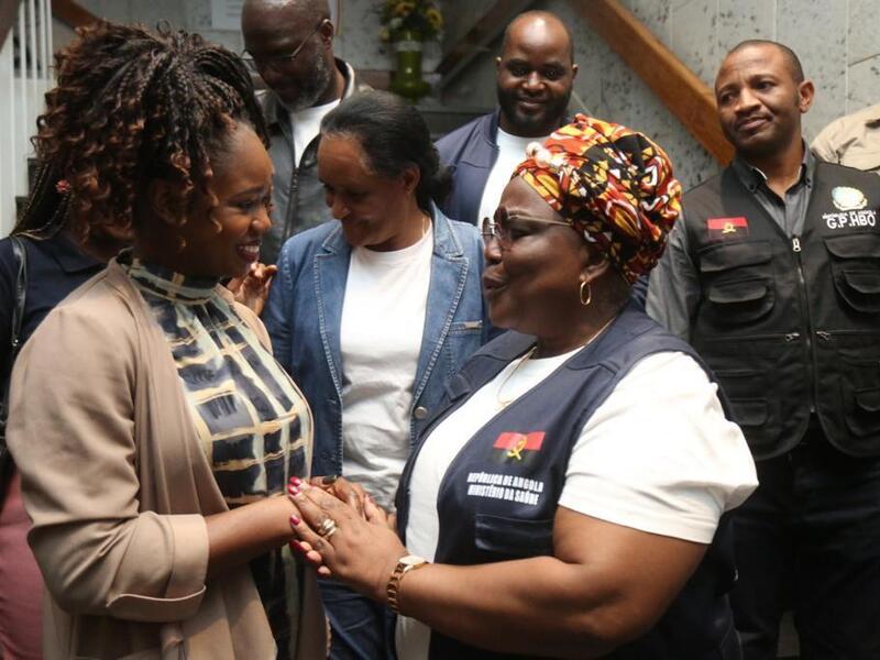 A photo of two women facing each other and smailing. The woman on the right is holding the women on the left's hands. Behind them is a crowd of people standing on a staircase. 