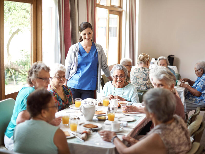 A dining room with two tables that have older people eating at them. There is a younger woman wearing a nurse's scrubs watching the people eat and smiling. 