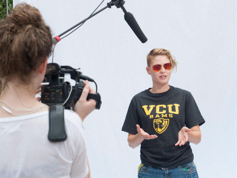 A photo of a woman speaking while standing in front of a white background. In front of her, a man with a microphone holds it above her head, a woman points a video camera at her and a man stands to the side. 