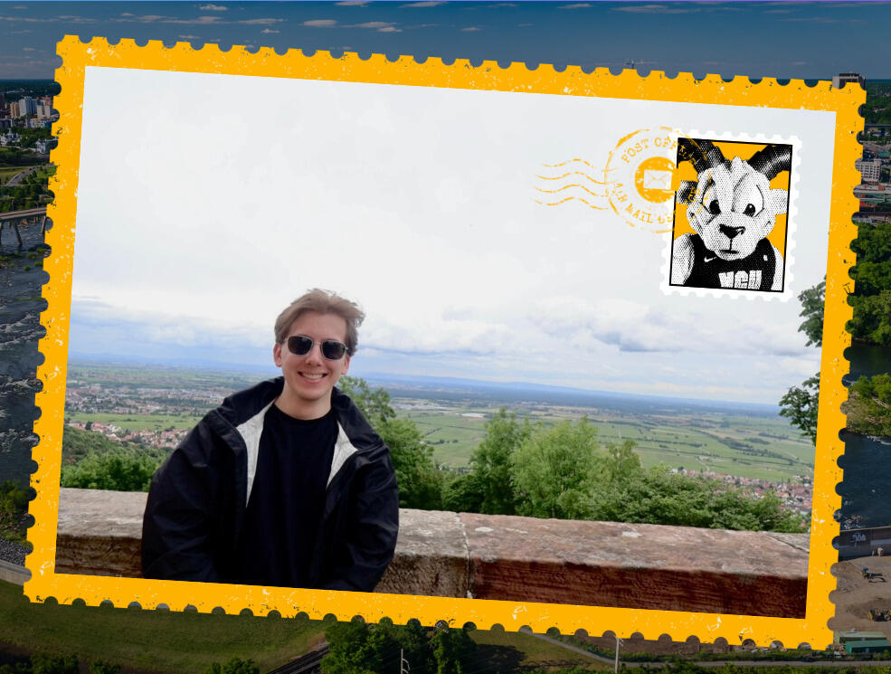 A photo of a postcard with a picture of a man standing in front of a short stone wall. Behind him are fields and a town. 