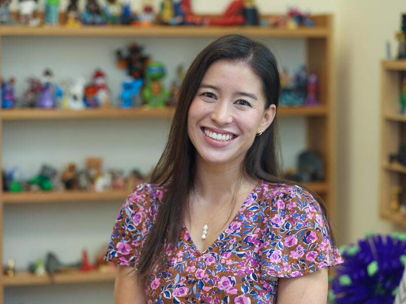 A photo of a woman from the chest up. She is sitting in front of a bookcase covered in toys. 