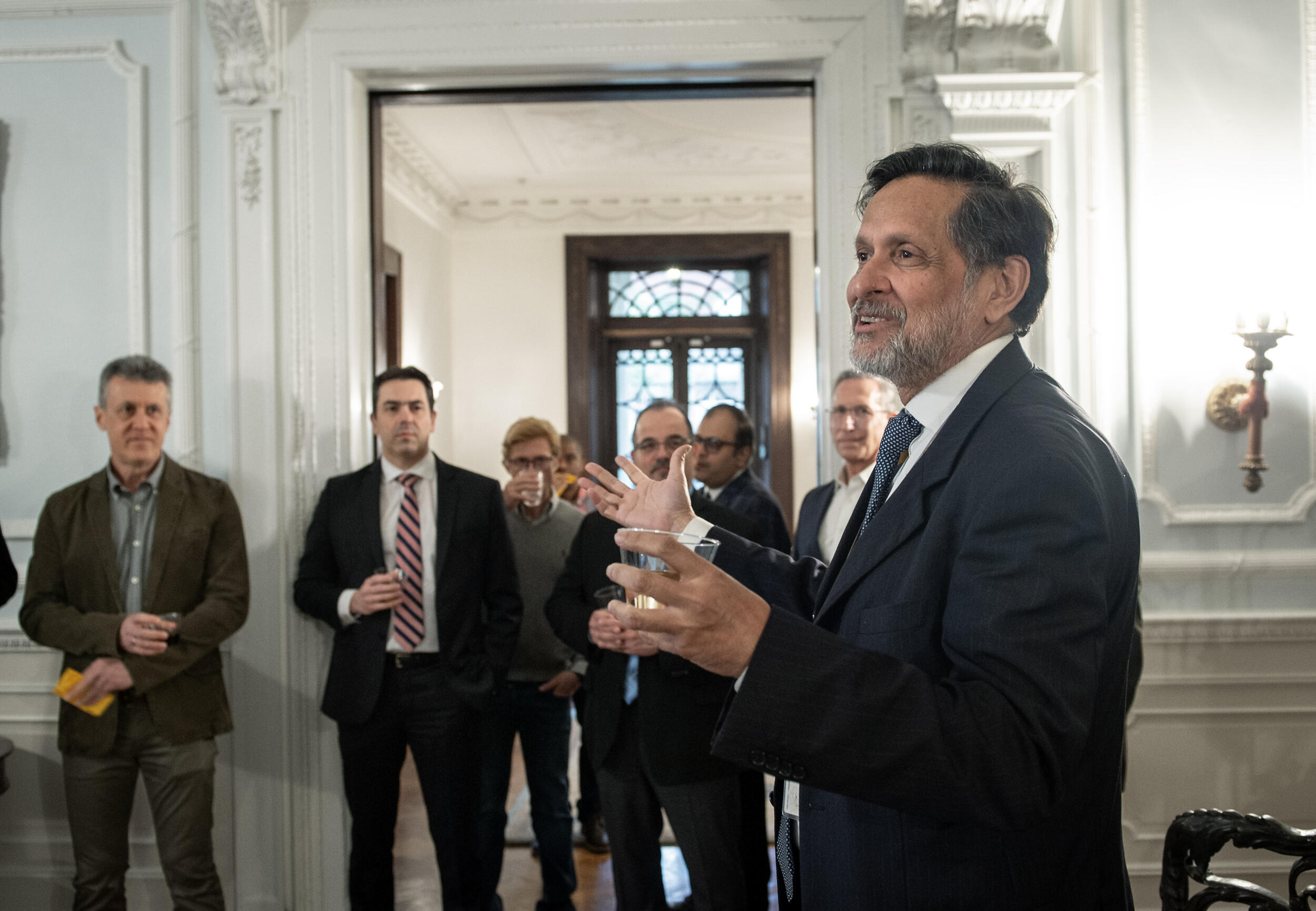 A photo of a man speaking while gesturing his hands. Seven people to his right are watching the man speak. 