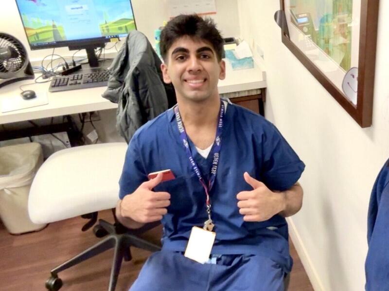 A man in blue hospital scrubs gives thumbs up, sitting in front of a computer screen.