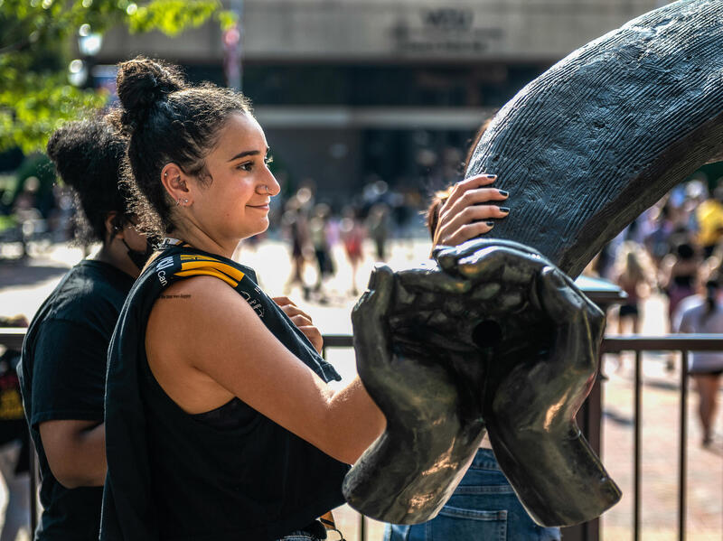 A student rests a hand on the Ram Horns outside The Commons in front of a plaza.