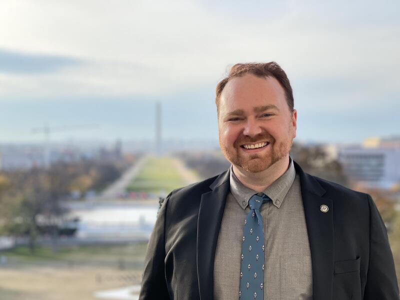 A photo of a man from the chest up. He is wearing a button up shirt, a tie, and a suit jacket. Behind the man is a blurry scenery of the National Mall in Washington D.C. 