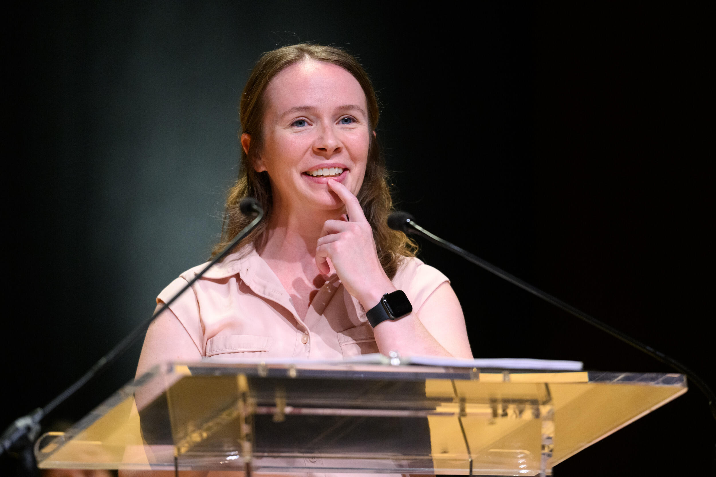 A woman behind a microphone and lectern smiles and holds a finger to her chin.