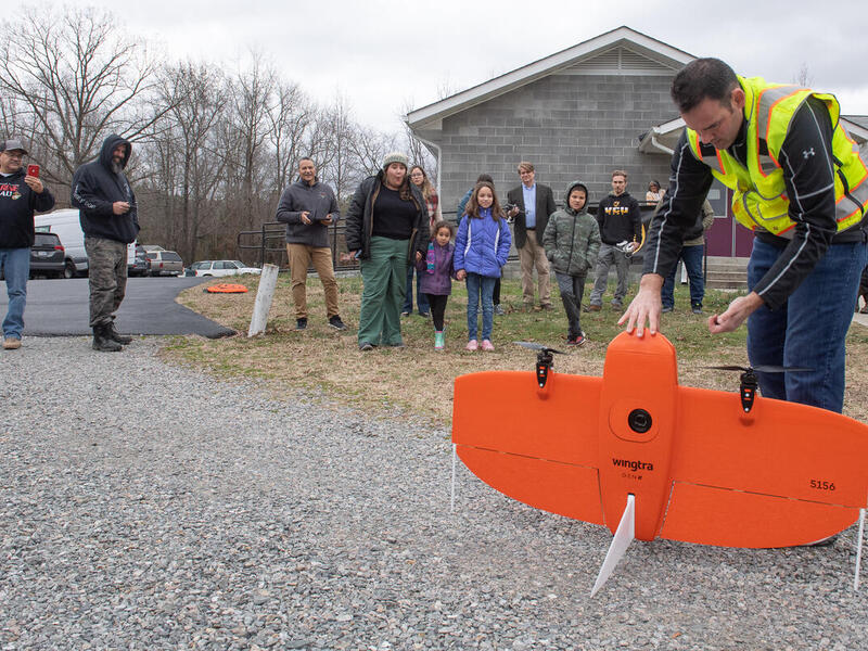 A photo of a man holding a large orange drone up as it balances on the ground. Behind him is a crowd of people watching him. 