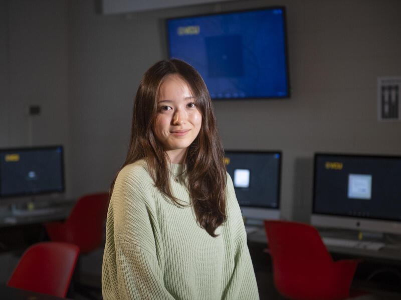 A photo of a woman from the waist up standing in a room with three computer monitors on desks, and red chairs.