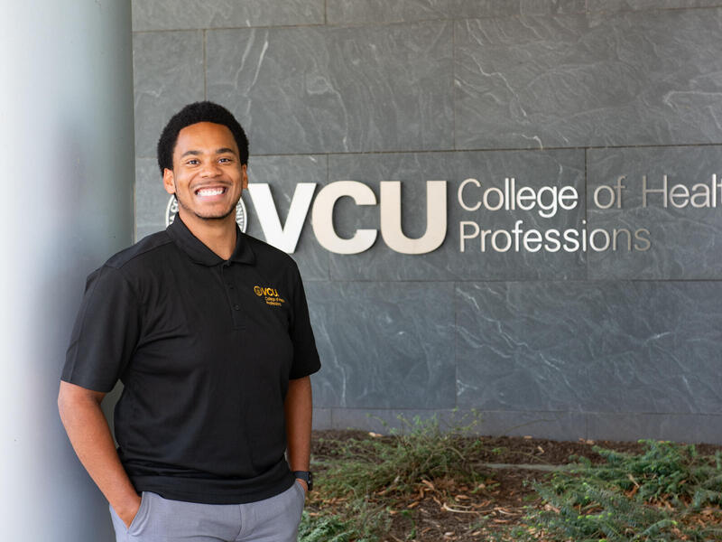 A photo of a man standing against a pillar and smiling. Behind him is a wall that says \"VCU College of Health Professions\" in silver letters.