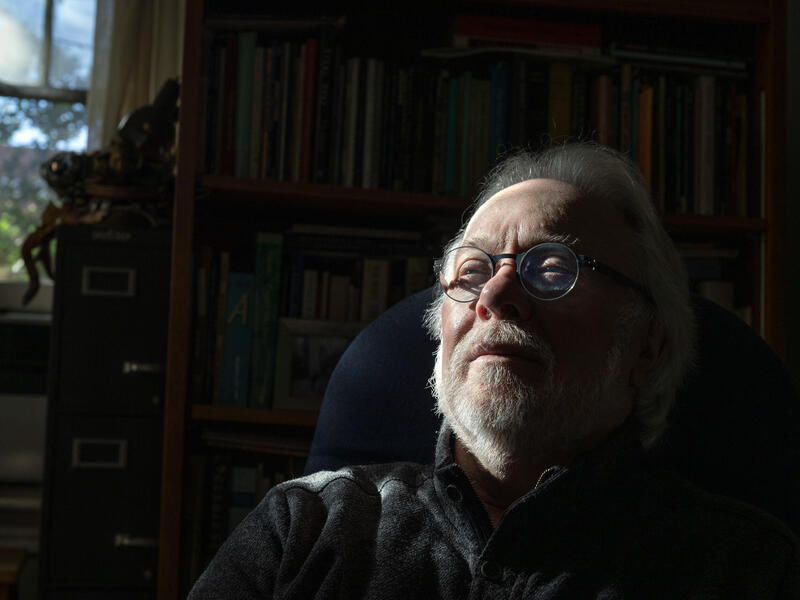A photo of a man sitting in a chair from the shoulders up. Behind him is a book case and a window. 