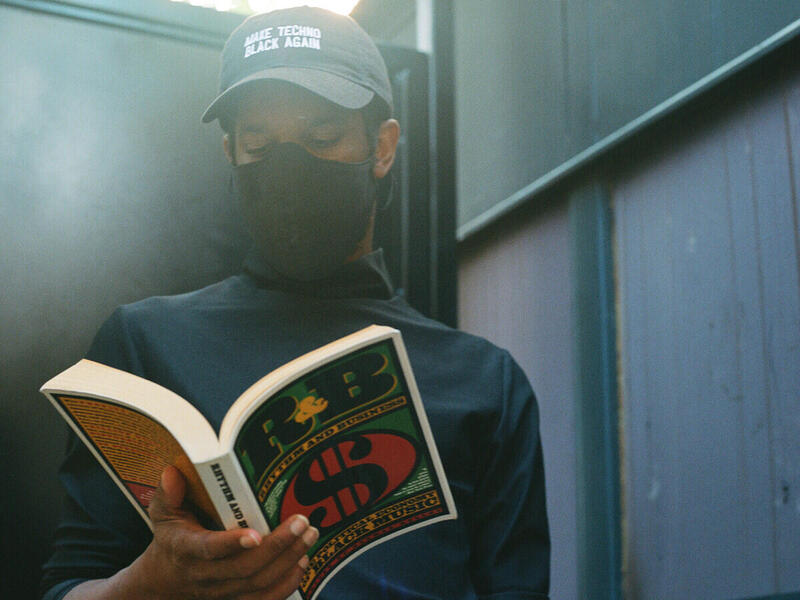 DeForrest Brown Jr reading a book while wearing a baseball cap and black face covering