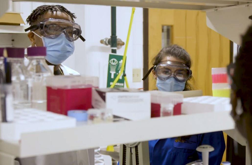 Two people wearing face masks, safety googles, and lab coats standing on the other side of a shelf filled with lab equipment. 