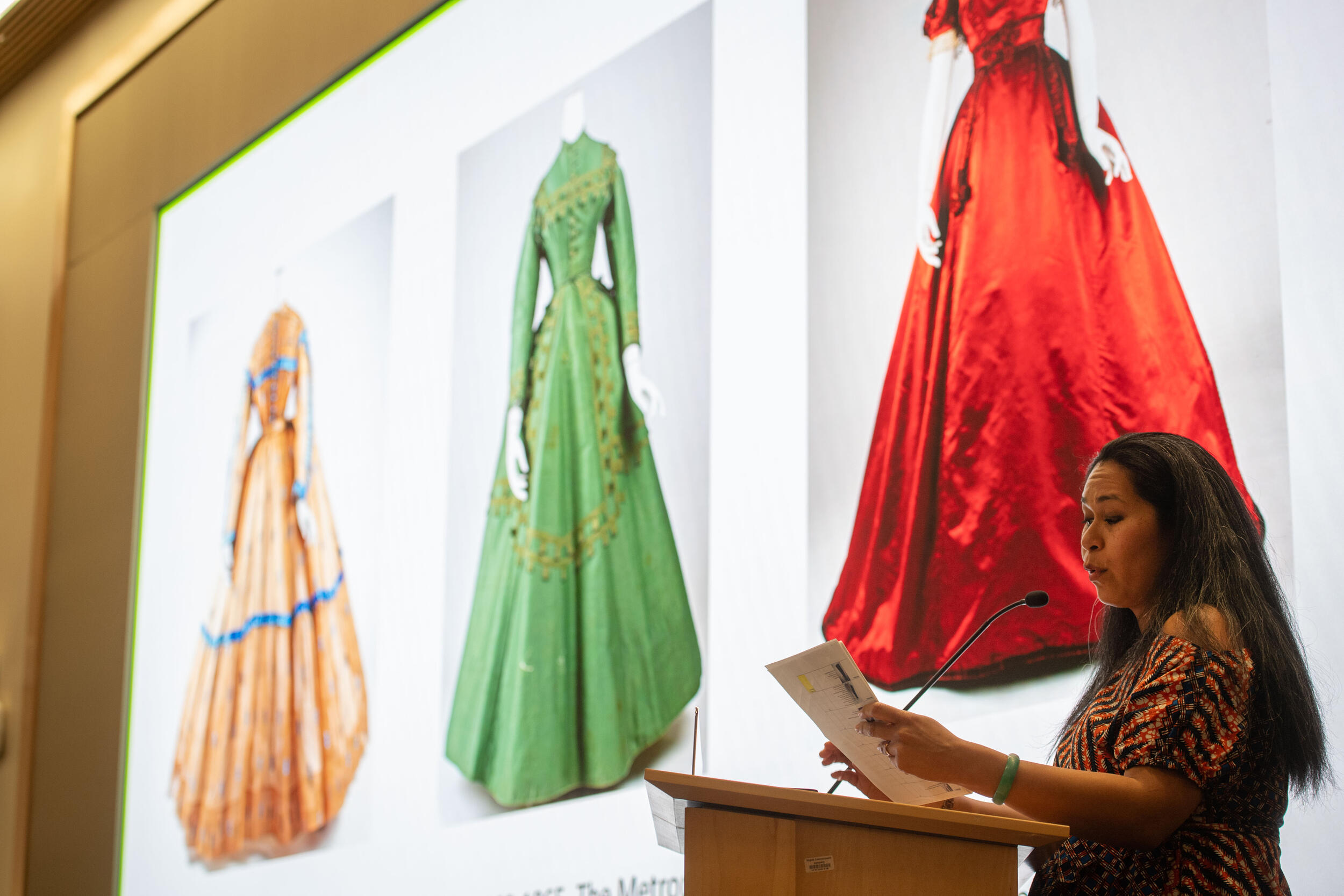 A photo of a woman speaking at a podium while holding a stack of papers. Behind her is a screen that shows three dresses from the 1800s which are cream, green, and red. 