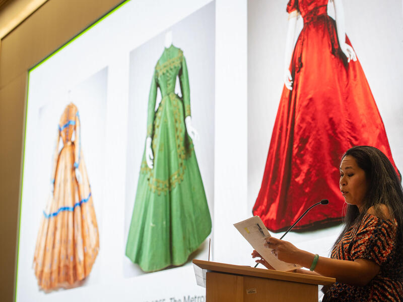 A photo of a woman speaking at a podium while holding a stack of papers. Behind her is a screen that shows three dresses from the 1800s which are cream, green, and red. 