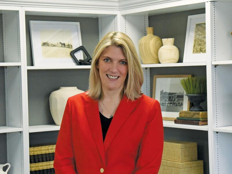 A portrait of a woman from the waist up wearing a red sports coat and black undershirt. She is standing in front of book shelves holding books, pottery, and framed pictures. 