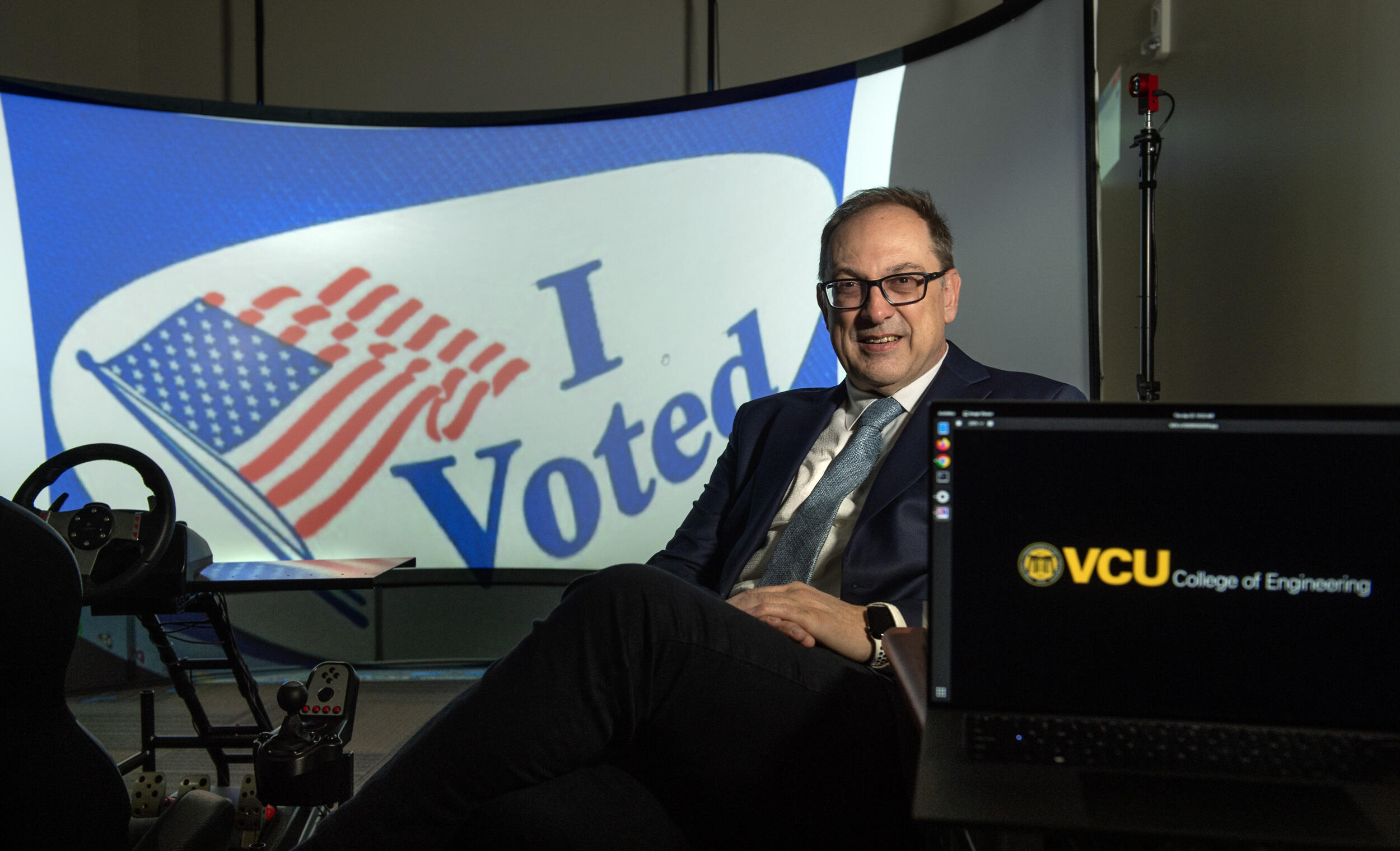 A man in a coat and tie sits in front of a screen with an \"I Voted\" message on it.