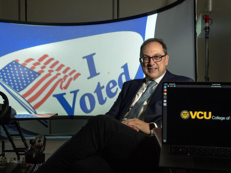 A man in a coat and tie sits in front of a screen with an \"I Voted\" message on it.