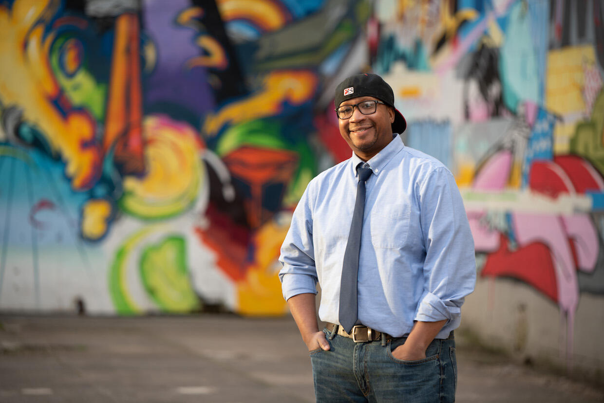 Marc Cheatham, wearing a backwards baseball cap, standing in front of a colorful mural.