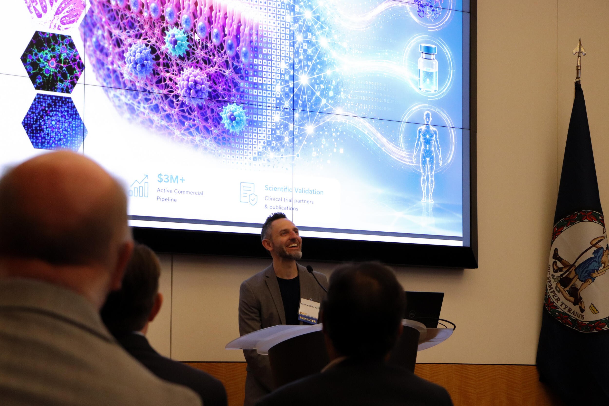 A photo of a man standing behind a podium speaking while people watch him. Behind the man is a screen that shows a 3D medical illustration of cells. 
