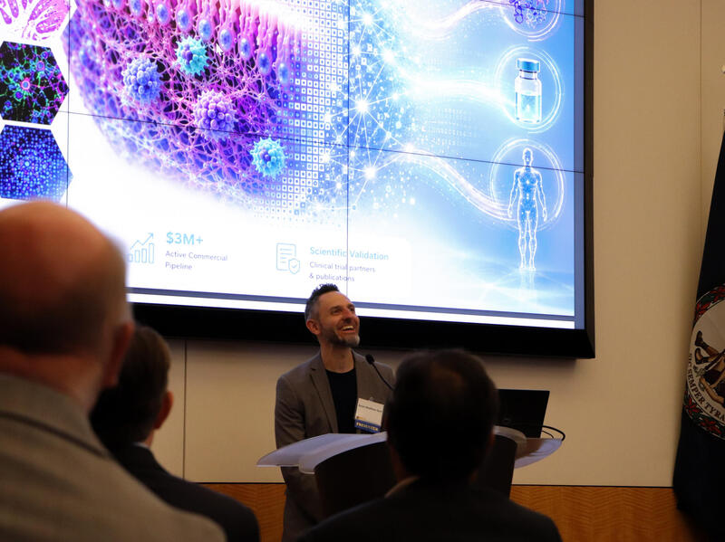 A photo of a man standing behind a podium speaking while people watch him. Behind the man is a screen that shows a 3D medical illustration of cells. 