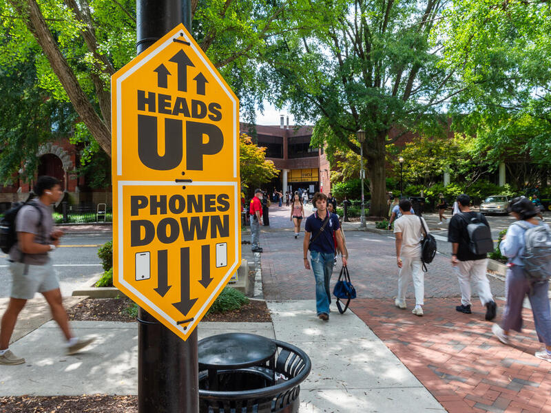 A photo of students walking across a crosswalk from a brick sidewalk to the Unvie4rsity Student Commons. On a pole next to the sidewalk is a large yellow sighn that reads &quot;HEADS UP&quot; with arrows pointing upwards and text that reads &quot;PHONES DOWN&quot; with an illustration of a smart phone and arrows pointing downwards.