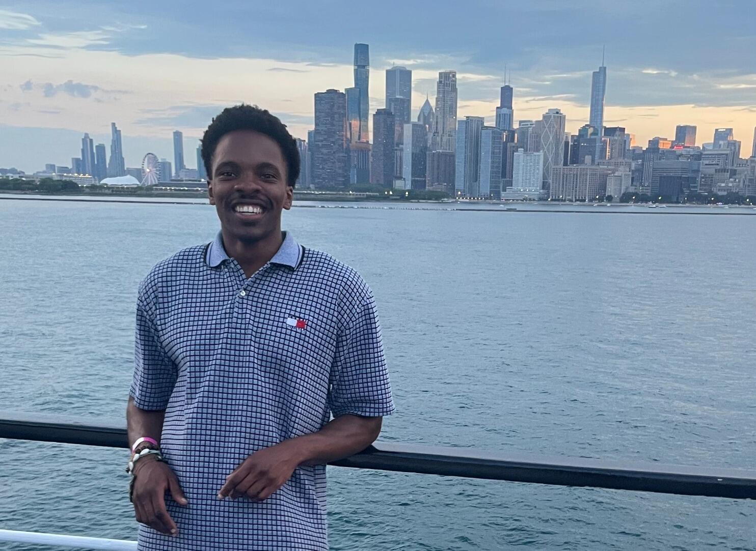A photo of a man standing in front of a railing seperating him and a large body of water. Behind the wter is a city skyline. 