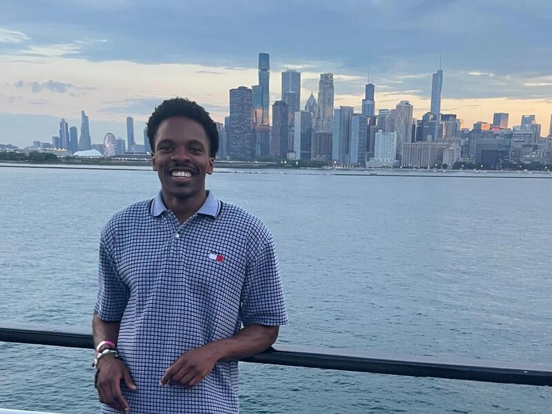 A photo of a man standing in front of a railing seperating him and a large body of water. Behind the wter is a city skyline. 