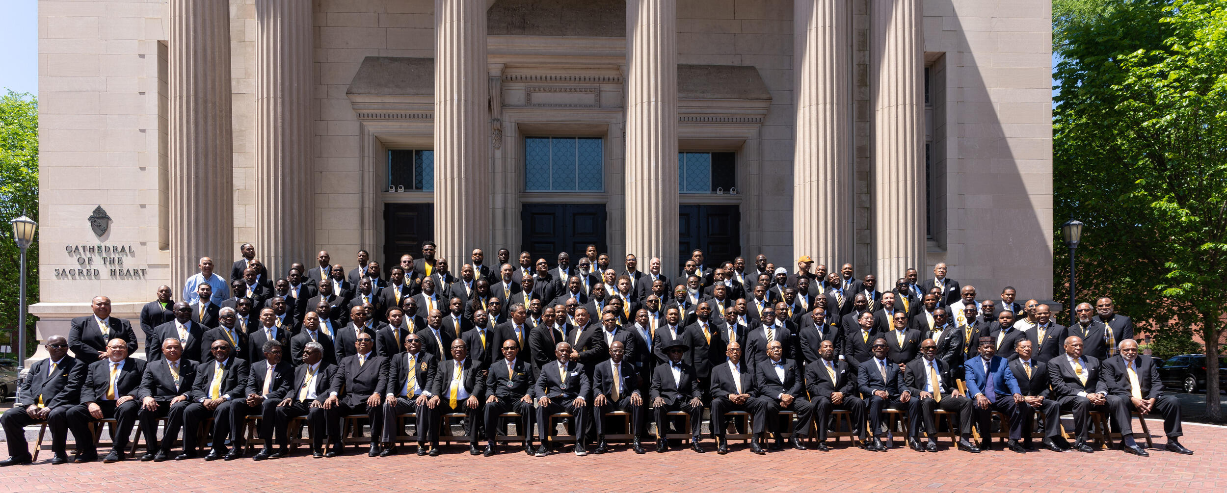 A large group of men sitting and standing in front of a chapel. 