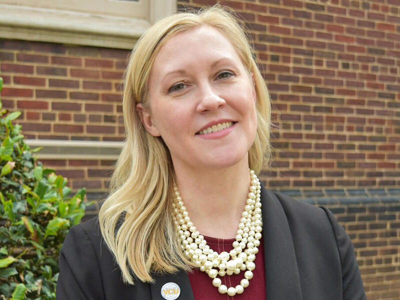 A woman standing outside a brick building next to a bush. 
