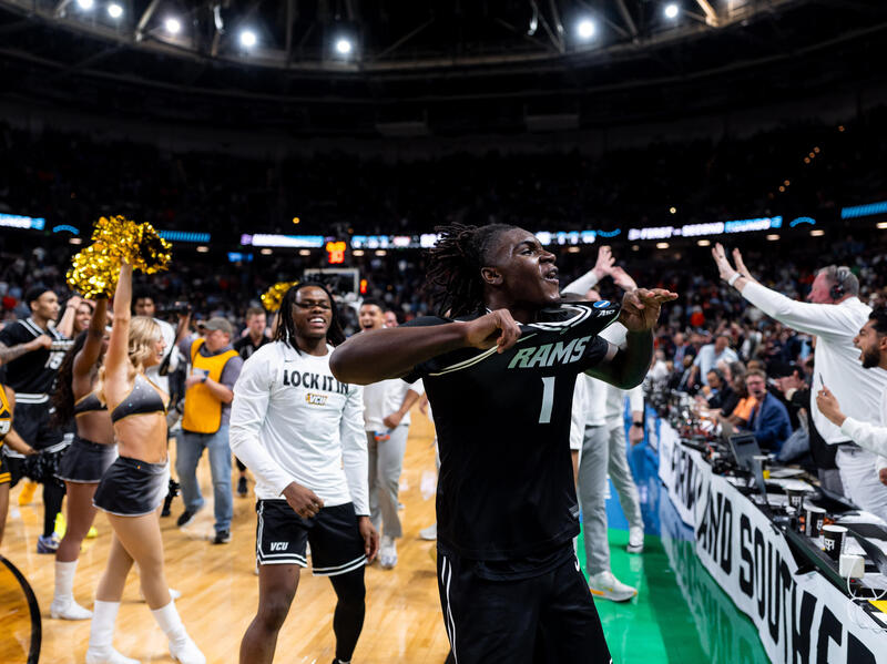 A photo of a man showing off his VCU Rams basketball jersey to a cheering crowd while standing on a court. 