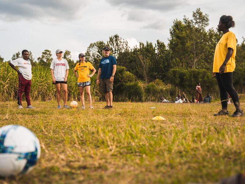 Center for Sport Leadership faculty and students stand in a soccer field along with local residents. A soccer ball is in the foreground.