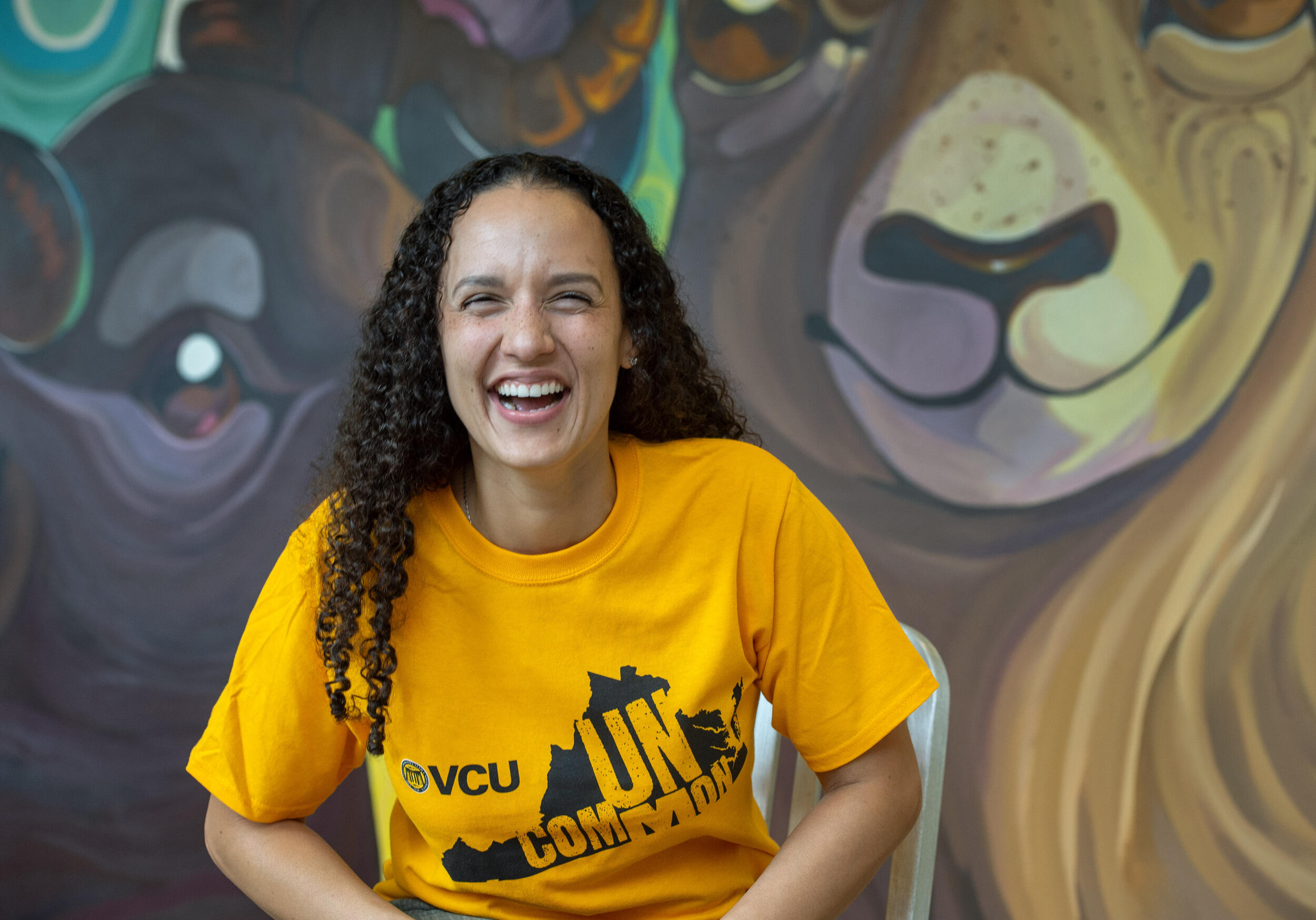 A photo of a woman laughing from the chest up. She is wearing a yellow t-shirt with the shape of Virginia printed on it. Inside the state's silhouette is yellow text that reads \"UNCOMMON.\" Behind the woman is a mural of two rams. 