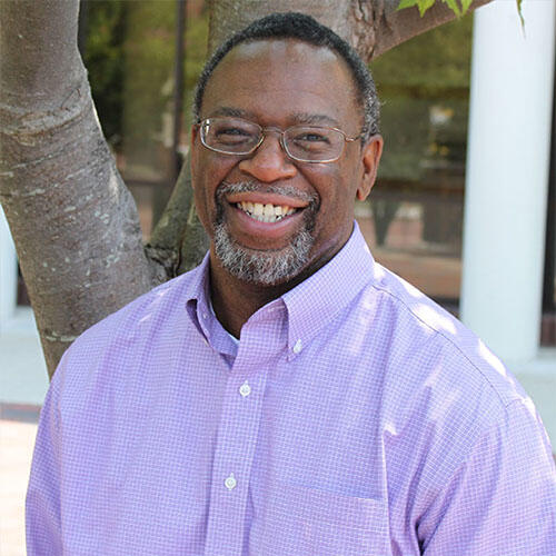 A man in glasses and a dress shirt smiles in front of a tree.