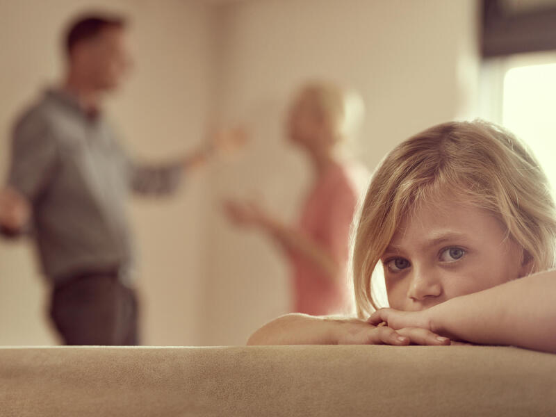 A child sadly leaning on a couch while in the background a blurry image of a woman and man look like they're fighting with each other. 