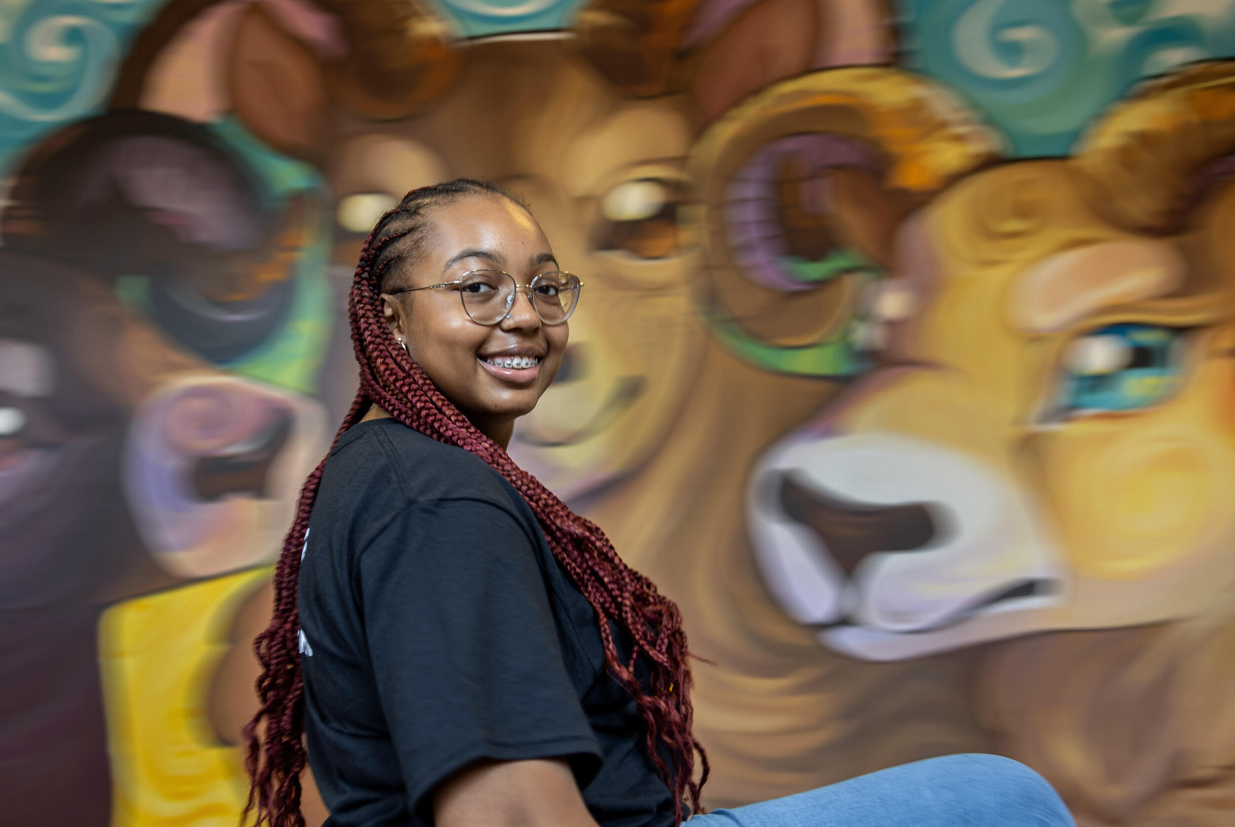 A photo of a woman from the chest up. Her body is turned away from the camera but she has turned her head so it is visable. She is wearing a black t-shirt and glasses. 
