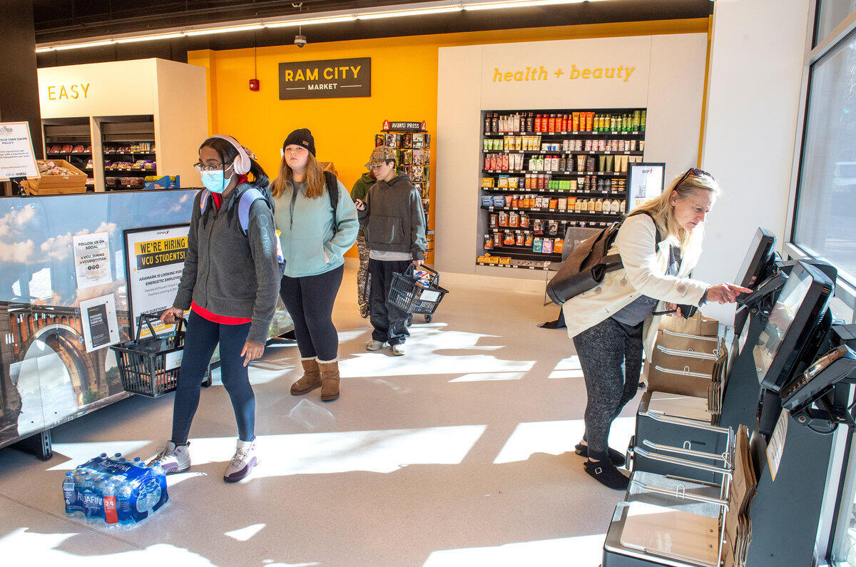 Three people lined up to the left and a woman on the right pressing buttons at a self-checkout machine. 