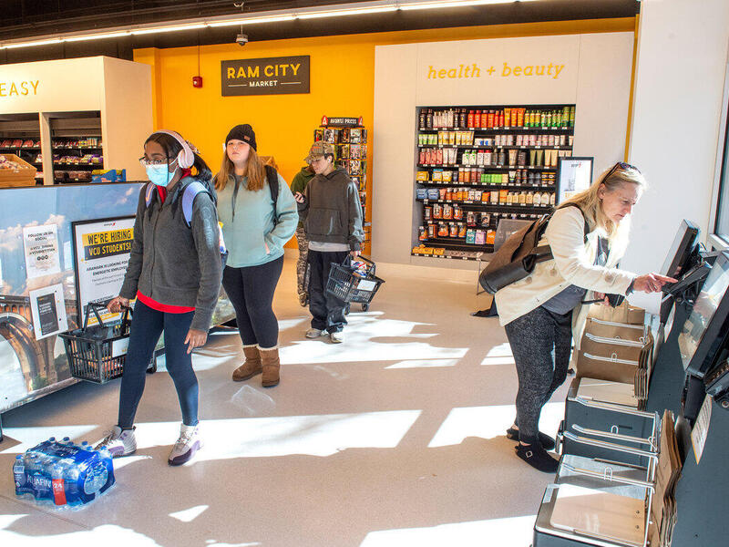 Three people lined up to the left and a woman on the right pressing buttons at a self-checkout machine. 