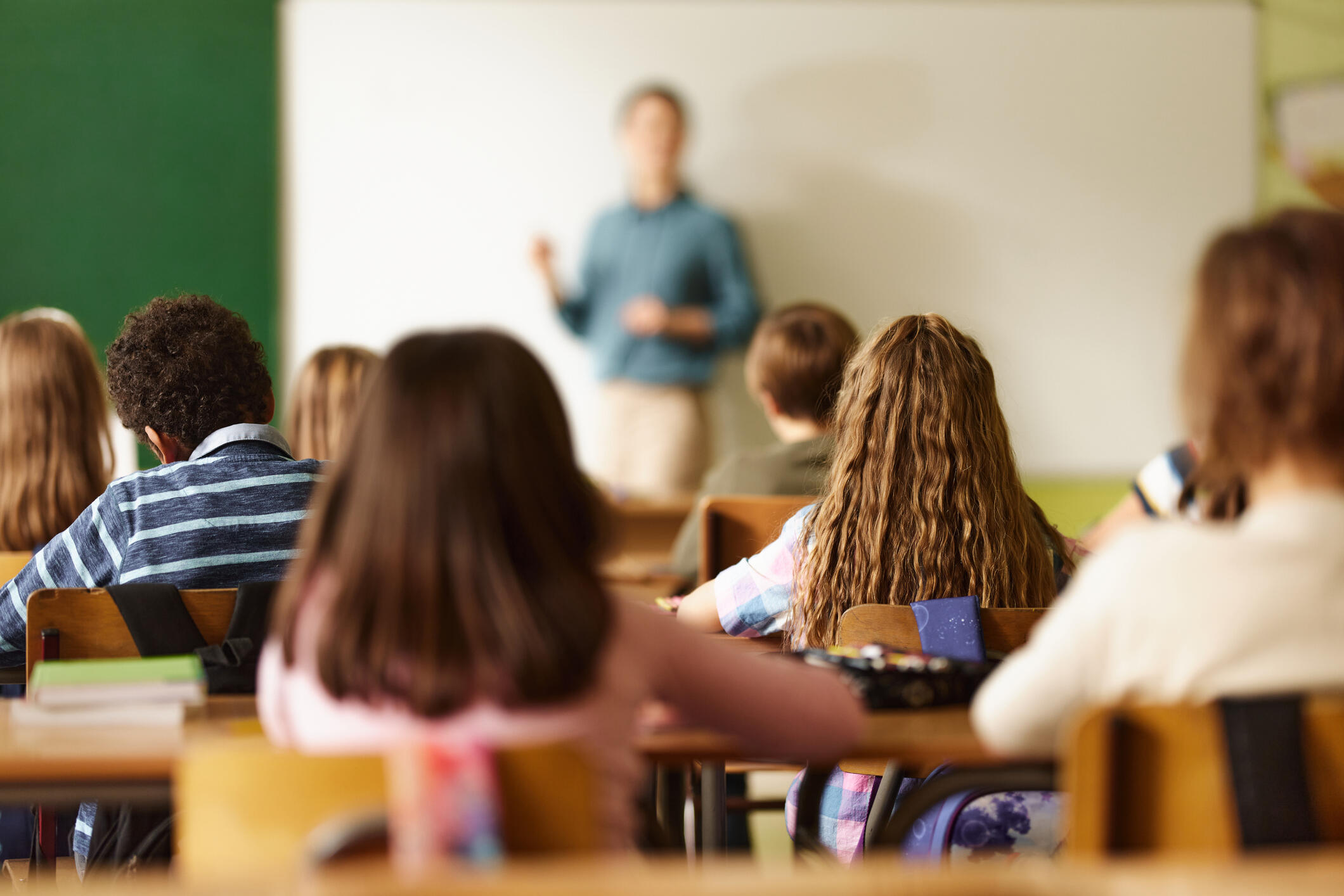 Children at desks in a classroom watch a teacher at the front of room talking.