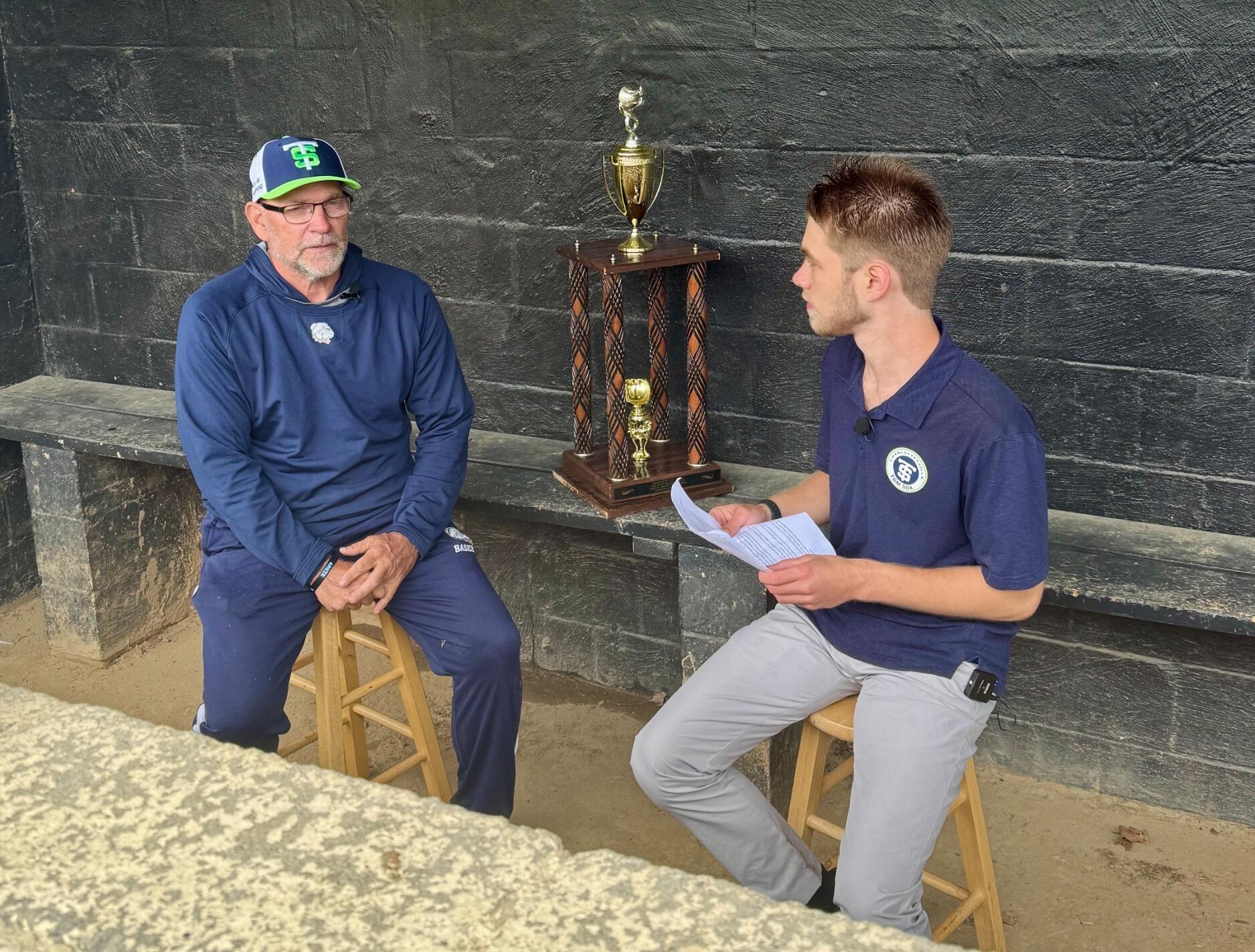 A photo of two men sitting on benches. The man on the right has a stack of paper in his hands. Behind the two men is a large trophy. 