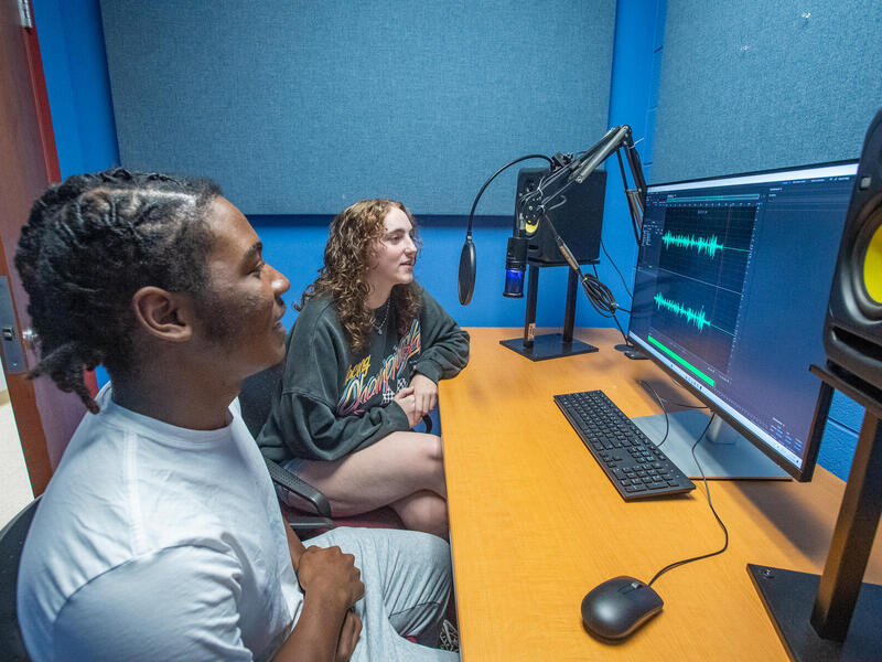 A photo of two people sitting at a table with a microphone, computer monitor, computer mouse, keypad, and speakers on it,. 