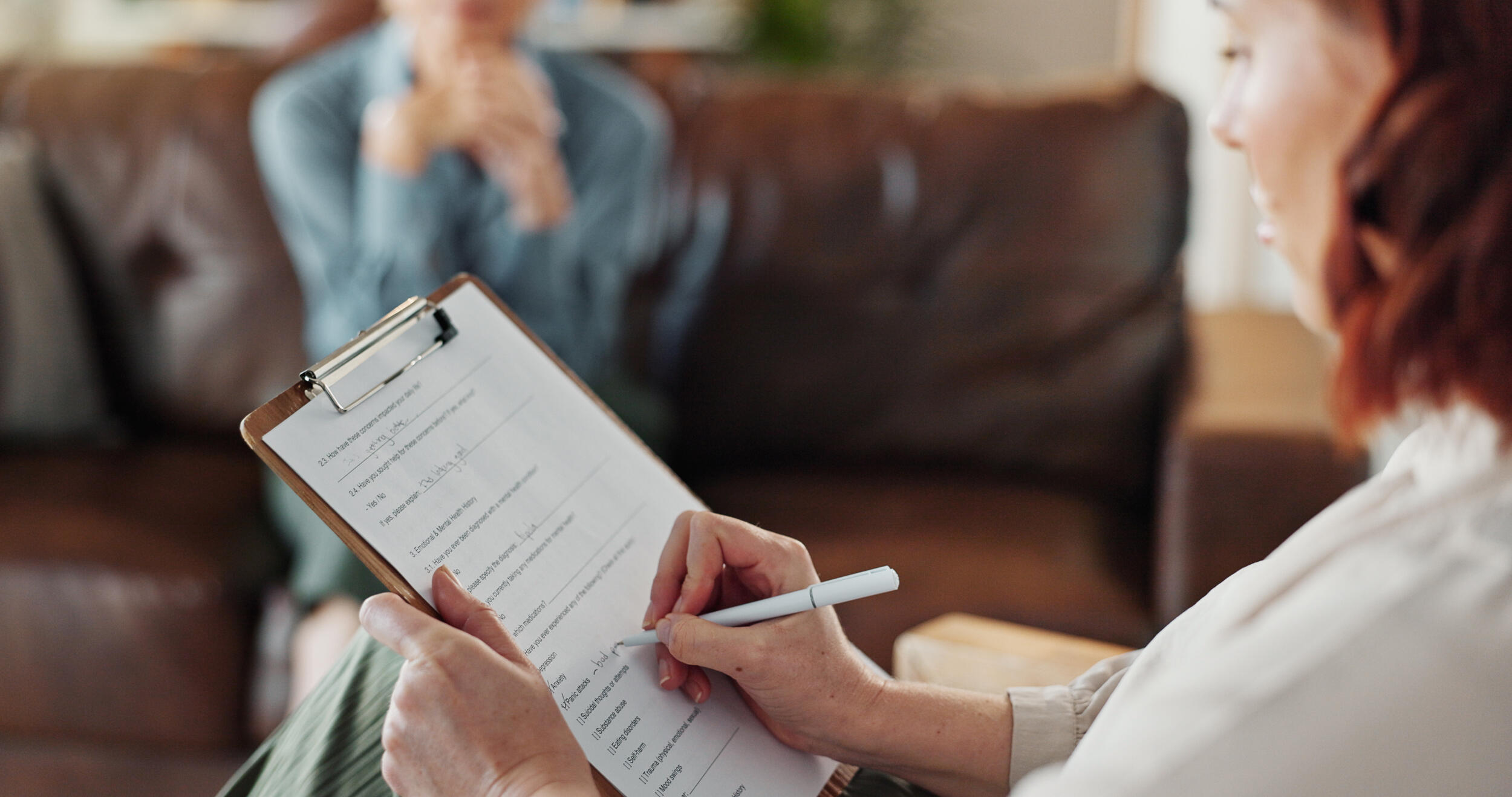 A photo of two people sitting in a room from across from eachother. One person is sittin on a couch, and the other is sitting in a chair and holding a pen and clip board. 