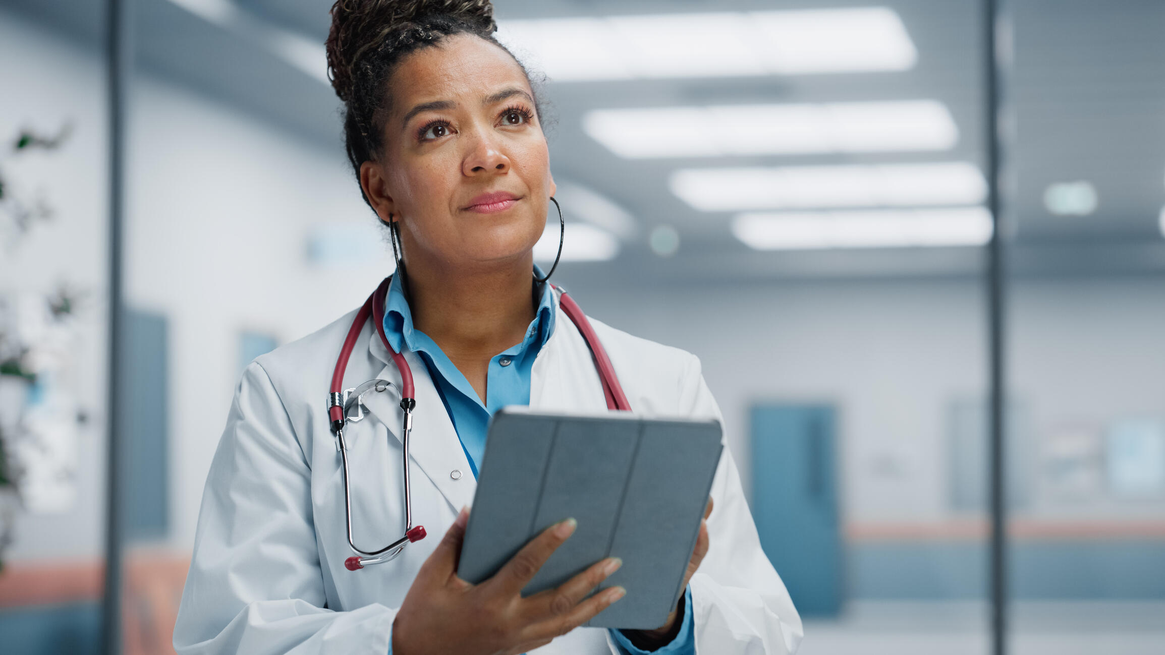 A photo of a doctor wearing a white lab coat and a stethoscope around their neck. They are holding at tablet and looking up. 