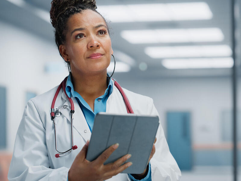 A photo of a doctor wearing a white lab coat and a stethoscope around their neck. They are holding at tablet and looking up. 