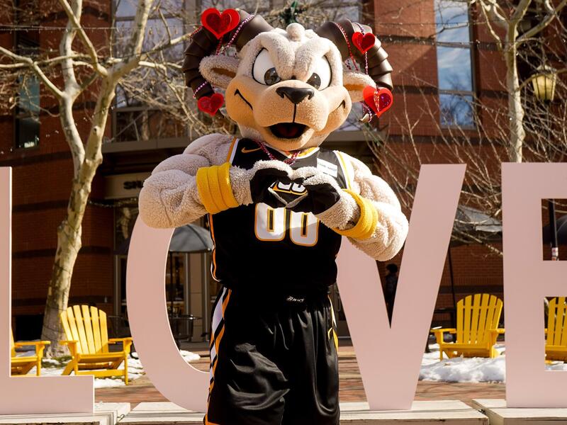 Mascot Rodney the Ram makes a heart shape with his hands, and stands in front of giant letters that spell love. His horns are decorated with hearts.