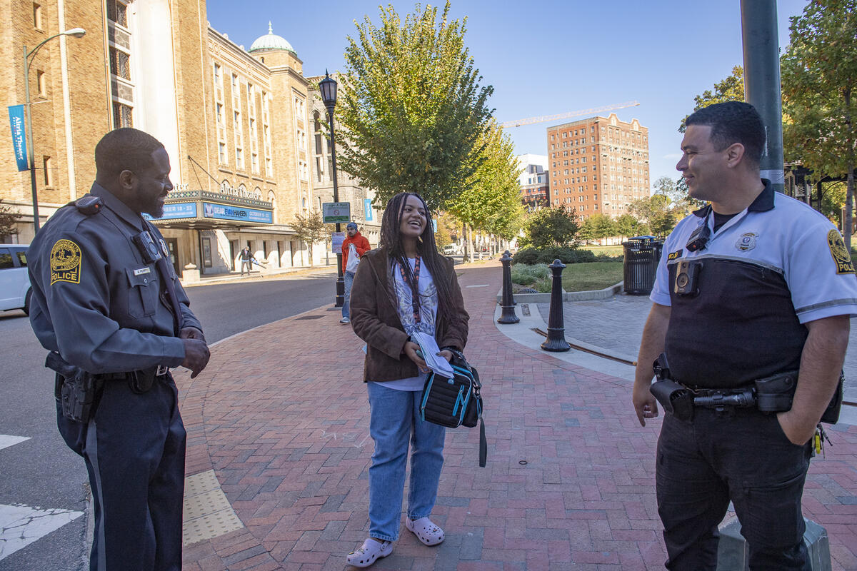 A woman standing between to VCU Police Officers on a sidewalk next to a park. 